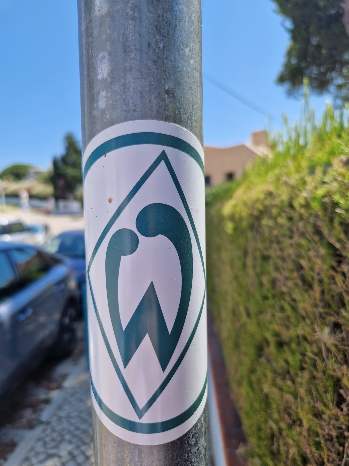 A dark teal and white diamond-shaped sticker with an abstract logo. The logo appears to incorporate the letter 'W' or a similar shape. The sticker is affixed to a metal pole outdoors.