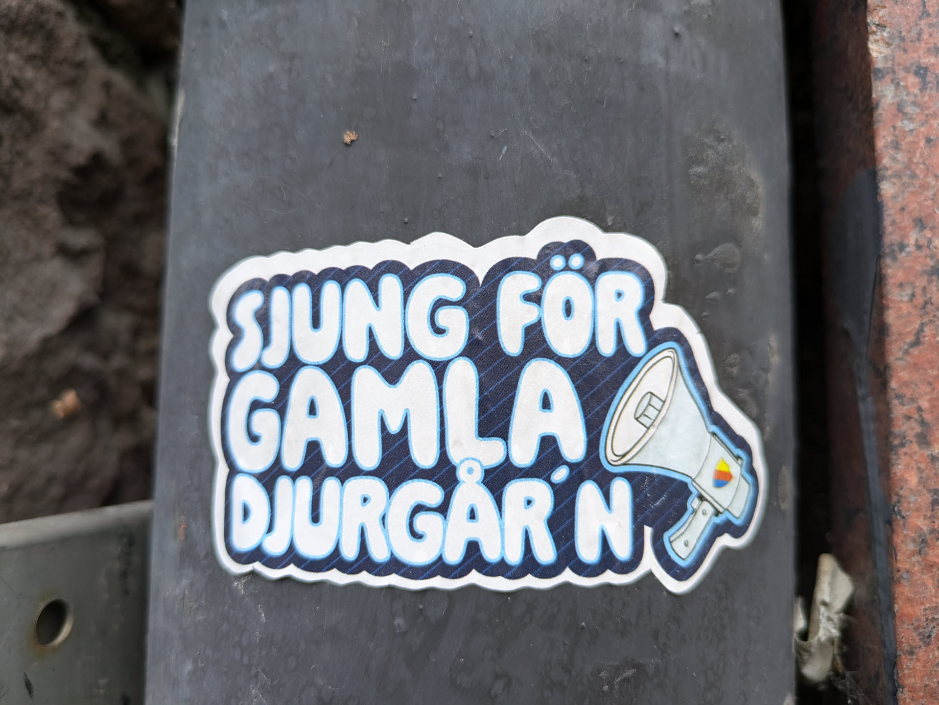 A sticker with the text "SJUNG FÖR GAMLA DJURGÅRDEN" in stylized lettering and an illustration of a megaphone.  The text translates from Swedish to "Sing for Gamla Djurgården". The sticker has a dark blue background with a subtle diagonal line pattern. 