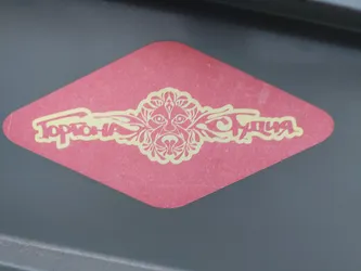 Red diamond-shaped sticker with a stylized floral design and the text "Topolina Studio" in a yellow-gold script. The design features a central face-like motif with ornate floral elements extending to either side.