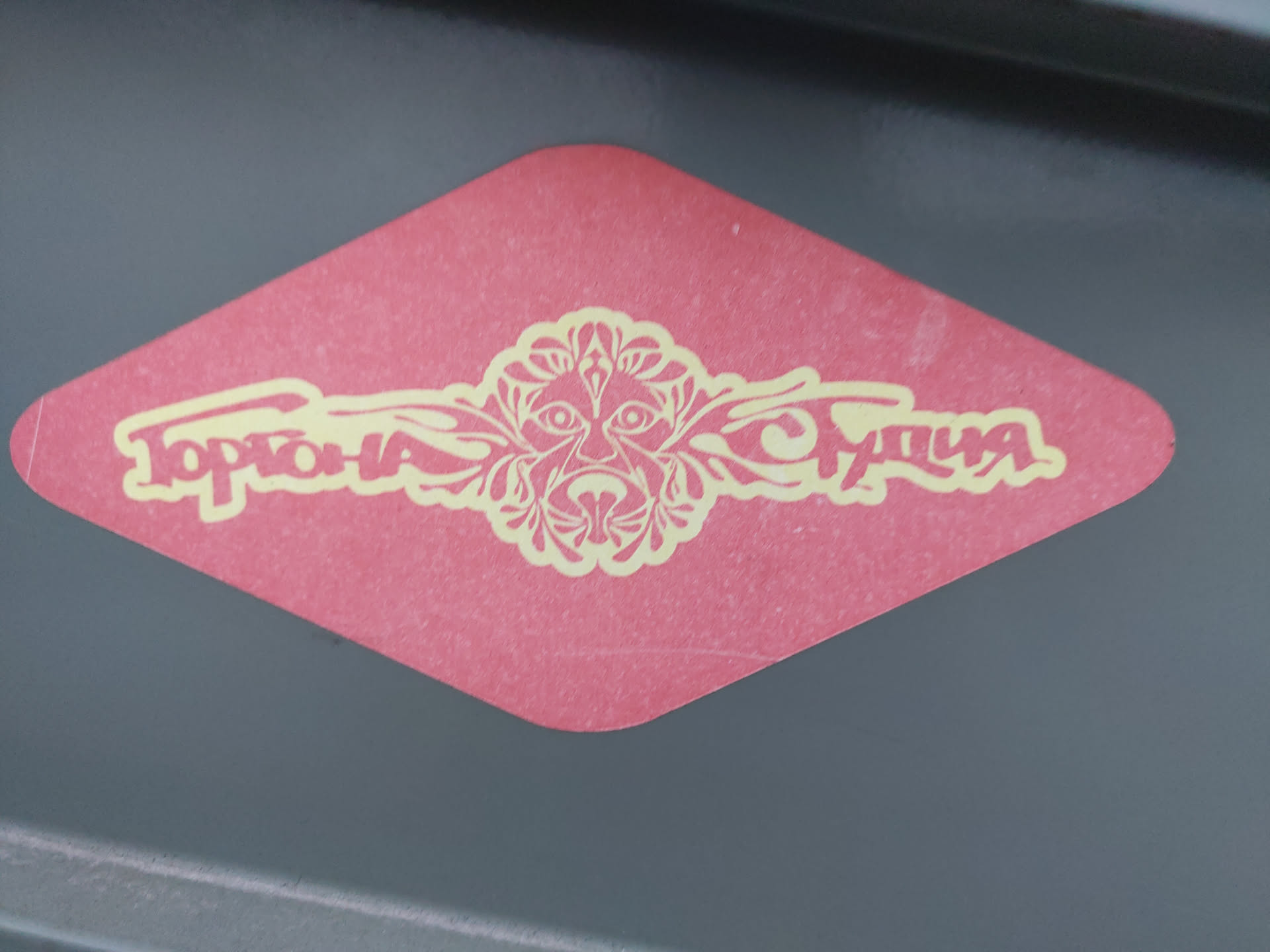 Red diamond-shaped sticker with a stylized floral design and the text "Topolina Studio" in a yellow-gold script. The design features a central face-like motif with ornate floral elements extending to either side.
