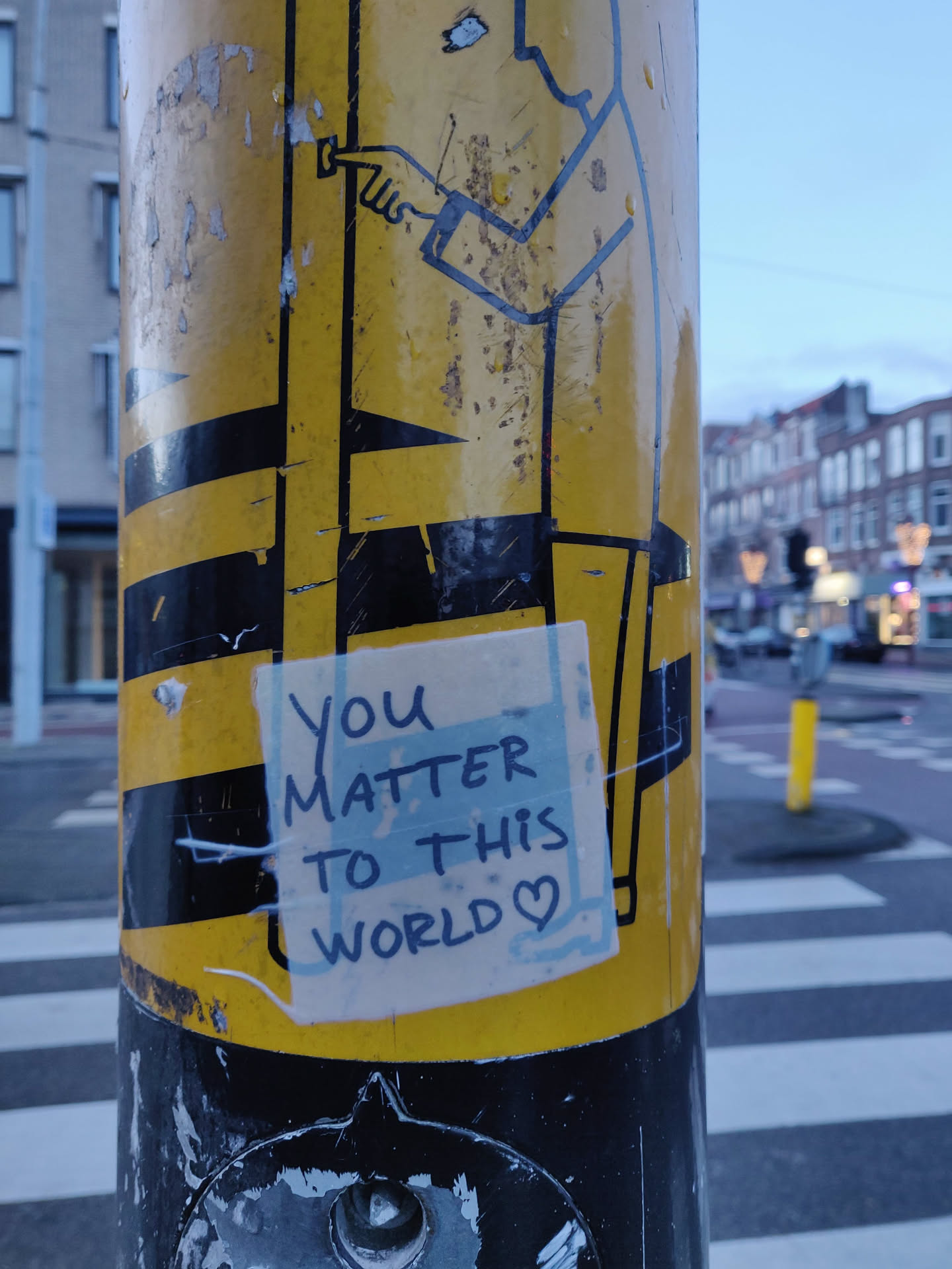 A small, yellow sticker with the text "You matter to this world" written on it in black ink and a small heart after the text. It's placed on a weathered yellow and black pole with other stickers and markings.
