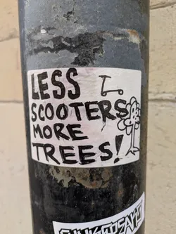 The image shows a sticker adhered to a pole. The sticker has a white background with black marker lettering. The text reads 'LESS SCOOTERS MORE TREES!' A simple line drawing of a scooter and a flower are adjacent to the text.