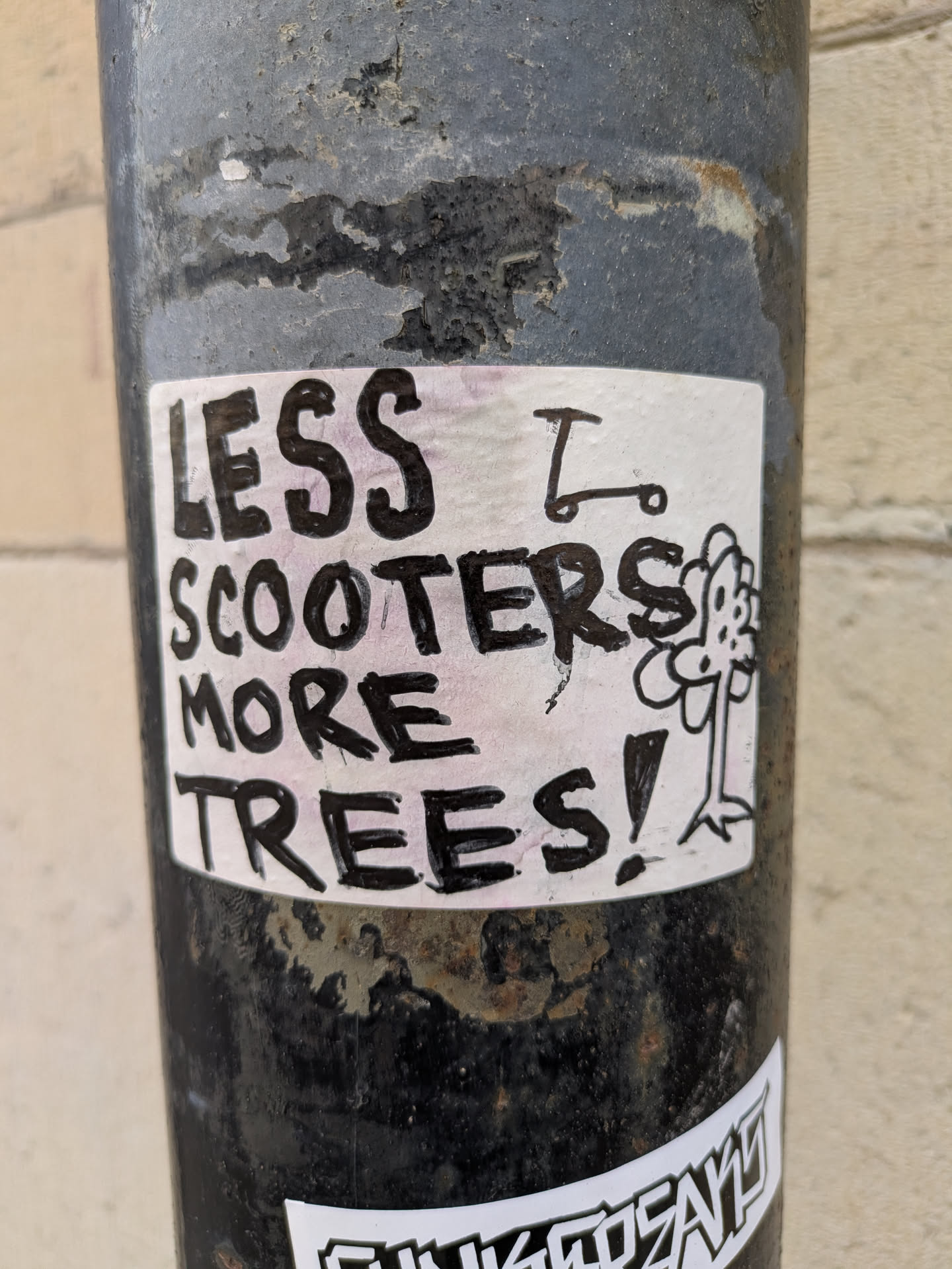 The image shows a sticker adhered to a pole. The sticker has a white background with black marker lettering. The text reads 'LESS SCOOTERS MORE TREES!' A simple line drawing of a scooter and a flower are adjacent to the text.