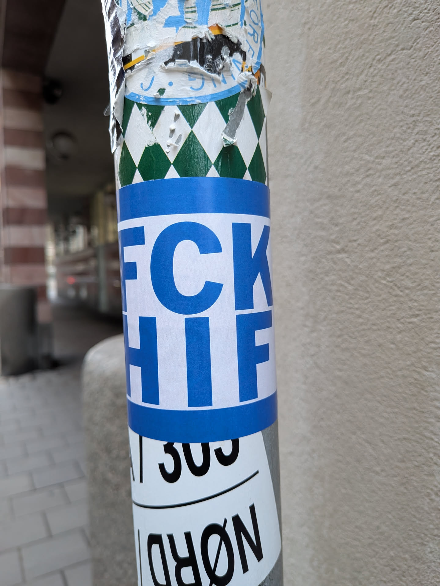 A rectangular sticker wrapped around a metal pole. The sticker features horizontal blue bands across the top and bottom, framing a white center. Bold blue sans-serif block lettering in the center displays 'FCK' on the upper line and 'HIF' on the lower line, commonly associated with football rivalry slogans against HIFK.