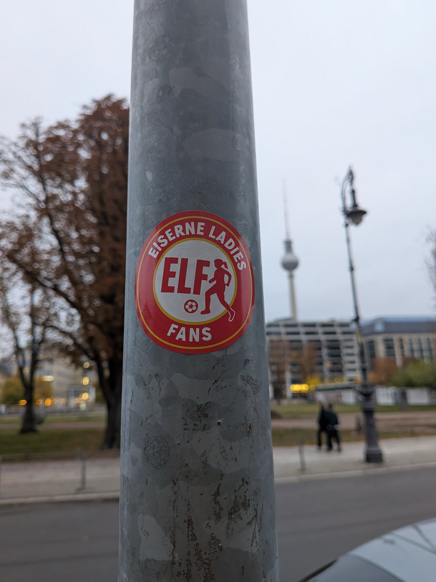 A circular sticker affixed to a metal lamppost. The sticker has a red outer ring bordered by yellow/orange trim, displaying the text 'EISERNE LADIES' along the top curve and 'FANS' along the bottom curve in white, bold, sans-serif letters. The inner circular field is cream-colored, featuring the bold red abbreviation 'ELF' next to a red silhouette of a female soccer player in mid-kick with a soccer ball underneath.