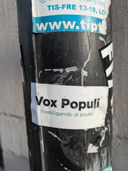 A white sticker with black text. It says "Vox Populi" and below in a smaller font "Investigando al poder". The sticker is on a dirty black pole.