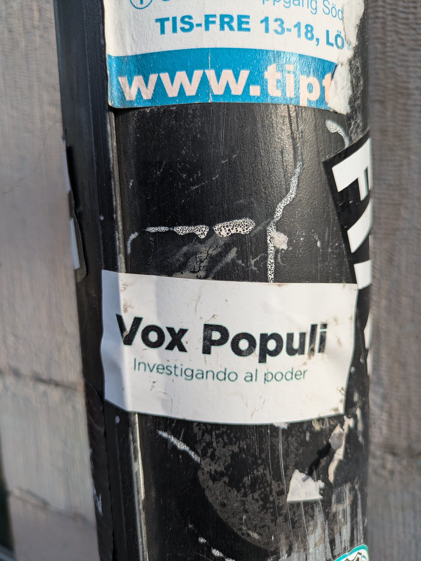 A white sticker with black text. It says "Vox Populi" and below in a smaller font "Investigando al poder". The sticker is on a dirty black pole.