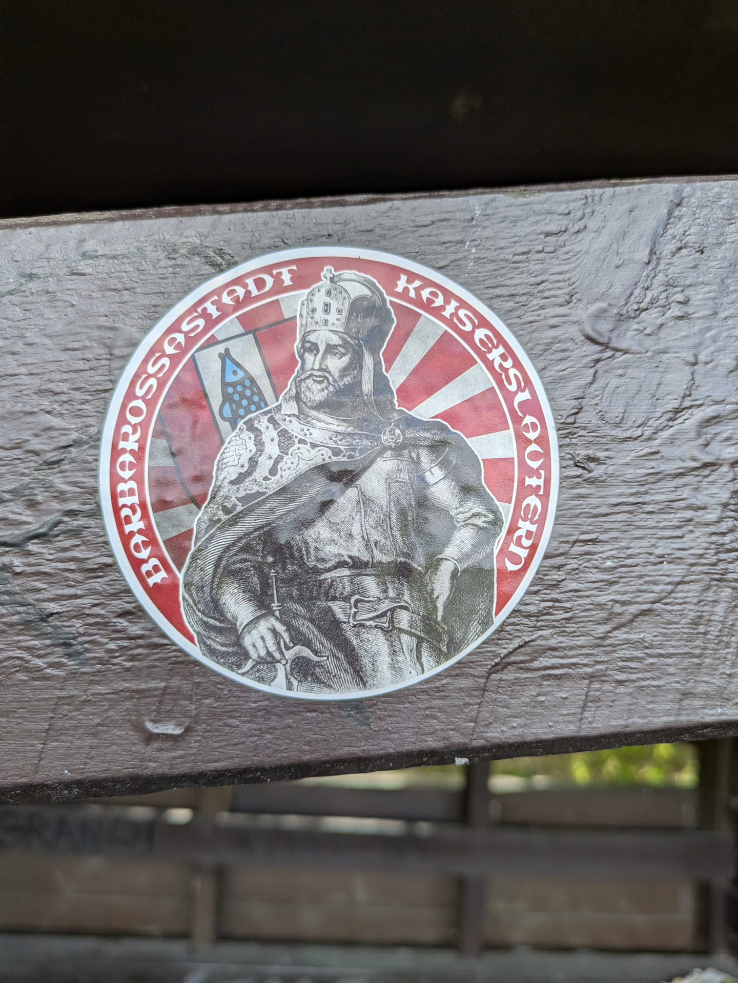 A round sticker features a depiction of Kaiser Barbarossa, the Holy Roman Emperor, against a red and white striped background. The sticker is placed on a wooden surface. Text around the image reads 'Barbarossastadt Kaiserslautern'.