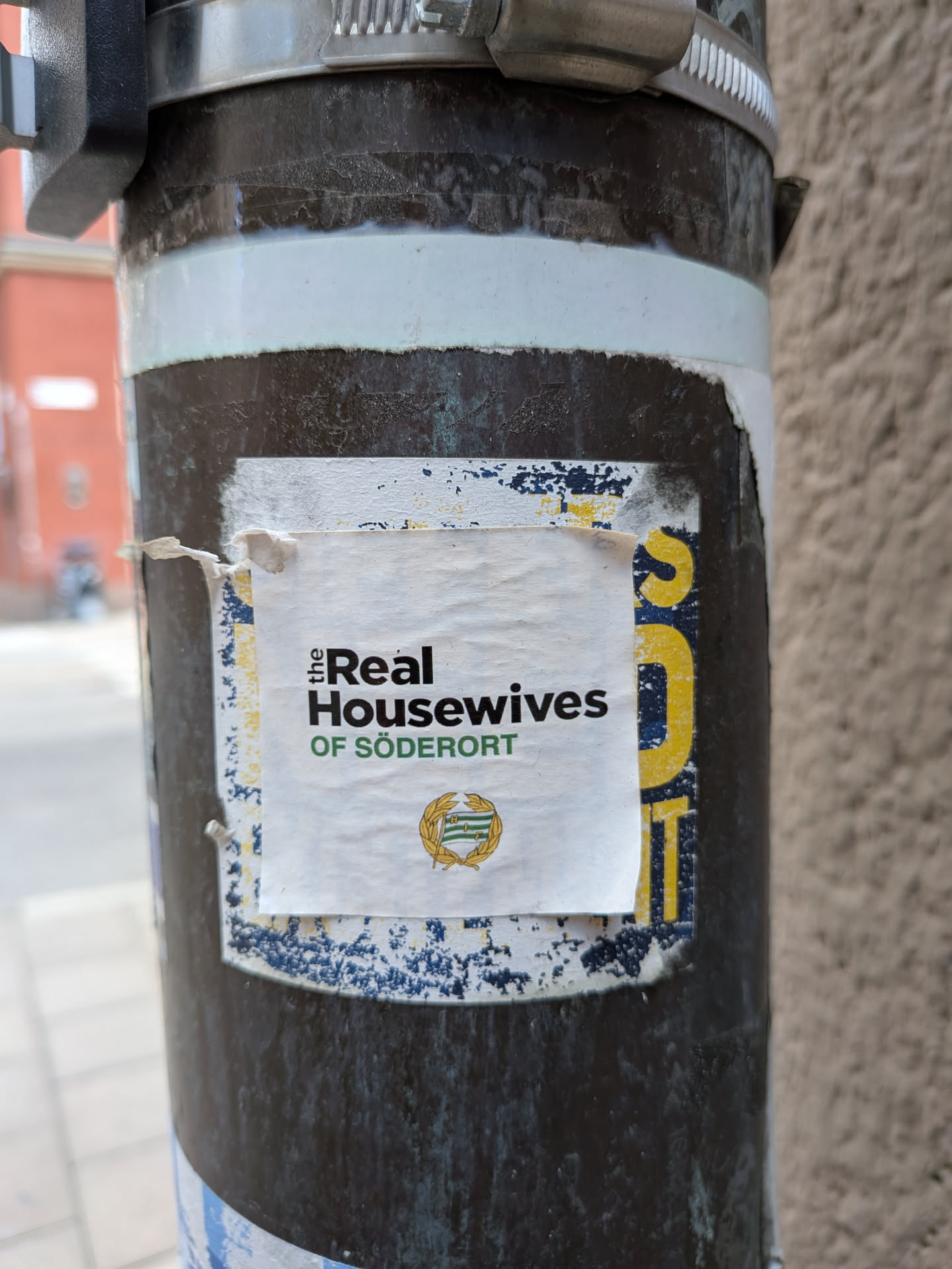 The sticker is white with black and green text. The top line reads 'the Real' in a serif font, with 'Real' being larger and bolder. Below that, in a sans-serif font, is 'Housewives'. The bottom line reads 'OF SÖDERORT' in green, also in a sans-serif font. In the lower center of the sticker is a circular emblem with a green and white design, possibly a coat of arms, surrounded by a gold wreath. The sticker is slightly creased and appears to be adhered to a dark, weathered pole, with a blue and yellow patterned layer underneath. There are also some blue and yellow paint splatters on the surface of the pole around the sticker.