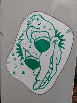 A green and white sticker featuring a whimsical creature. The creature appears to be a stylized dragon or similar mythical beast, with a friendly expression. The art style is simple and slightly rough around the edges, giving it a hand-drawn feel. The sticker is affixed to a grey surface, possibly a wall or a post.