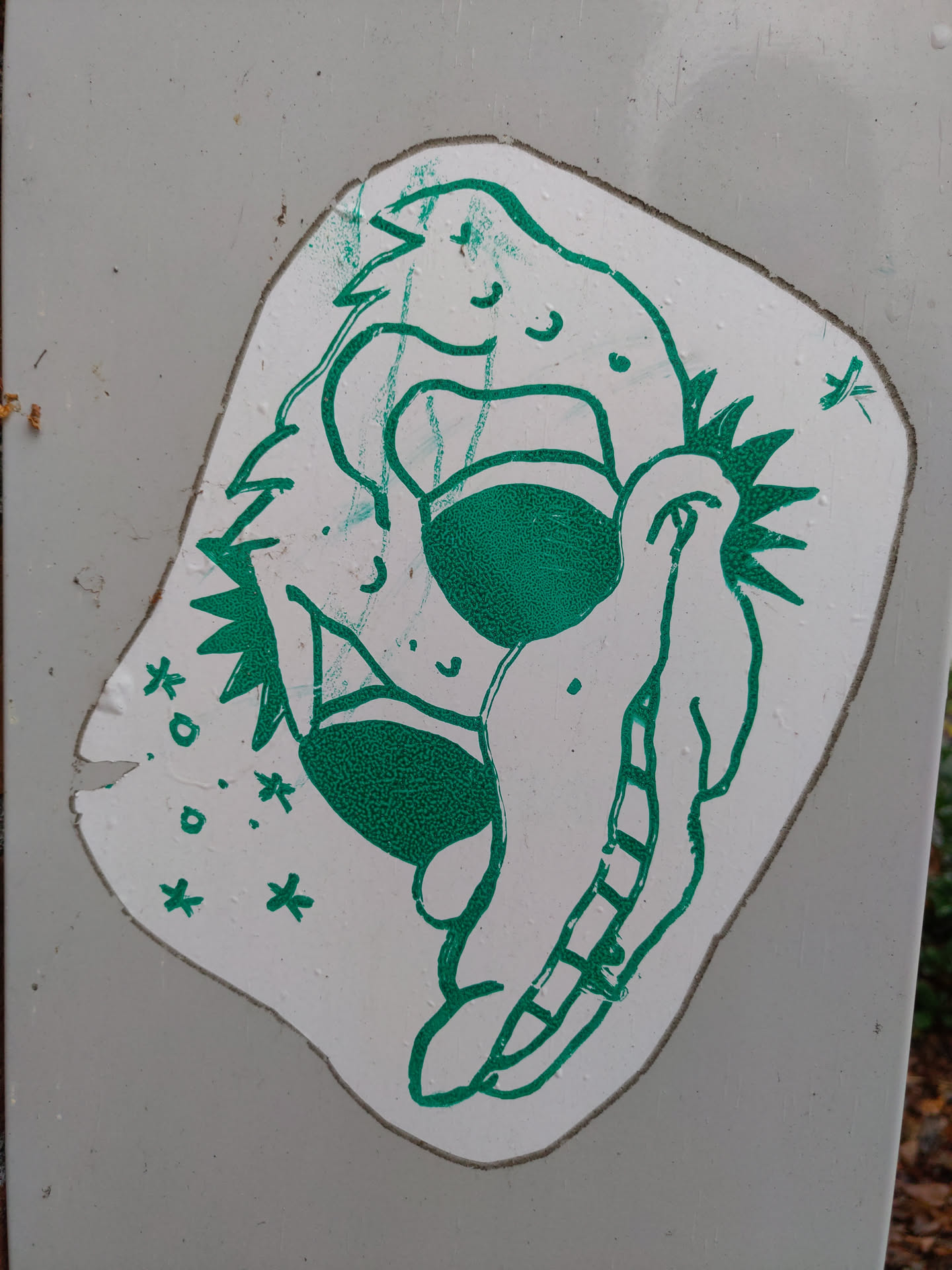 A green and white sticker featuring a whimsical creature. The creature appears to be a stylized dragon or similar mythical beast, with a friendly expression. The art style is simple and slightly rough around the edges, giving it a hand-drawn feel. The sticker is affixed to a grey surface, possibly a wall or a post.