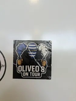 A square sticker with a black background. The image features a masked figure with blue and white striped face and yellow eyes, holding two flares that produce blue and white smoke. Below the figure, the text "OLIVEO 8 ON TOUR" is printed in a stylized font. The sticker appears to be placed on a white surface.