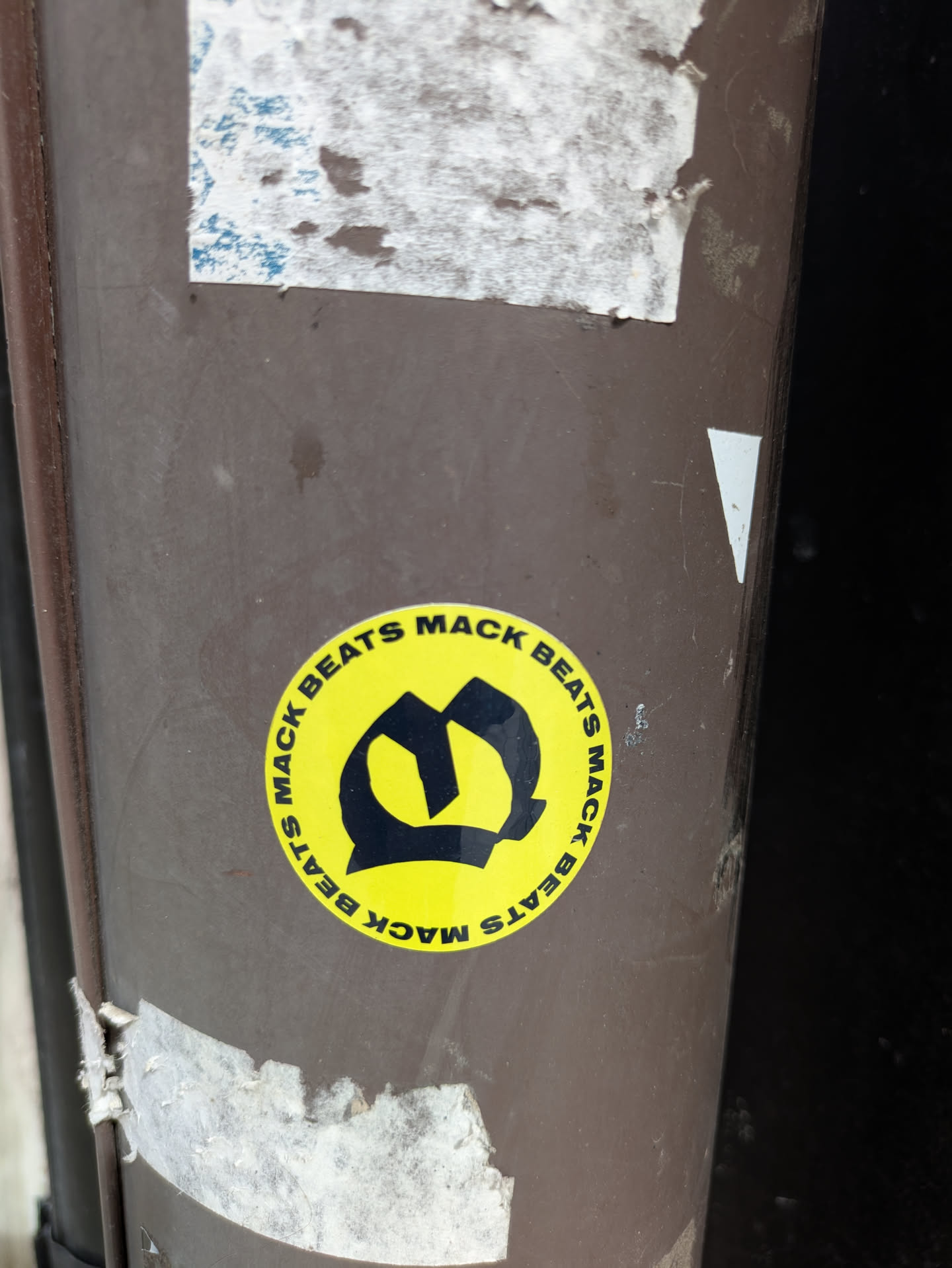 The image features a round, yellow sticker with the text 'MACK BEATS' circling a black, stylized letter 'M' in the center. The sticker is affixed to a brown, weathered surface, possibly a pole or post.