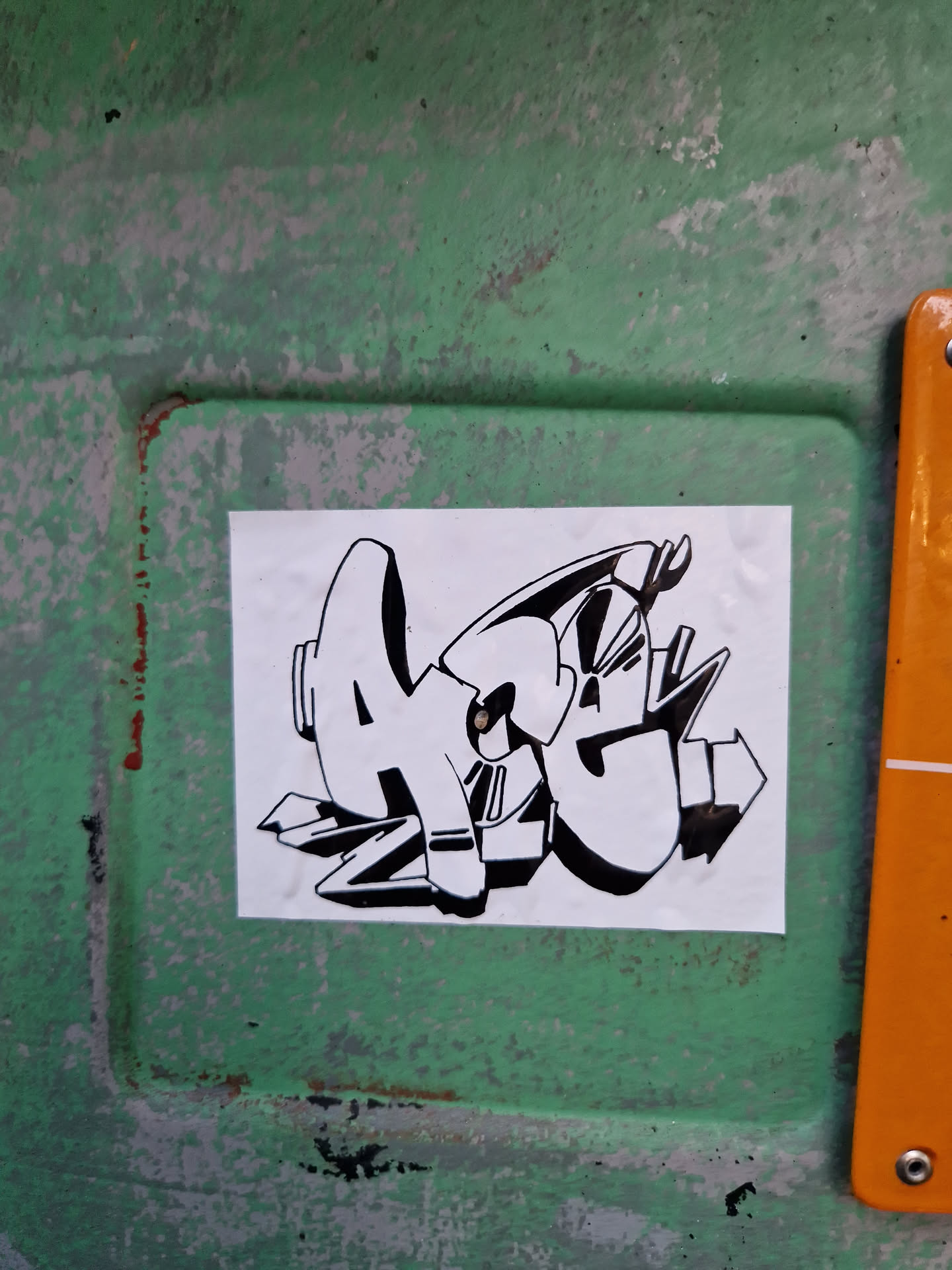 A square sticker with a black and white graffiti-style design. The graffiti appears to say "AGZ". The sticker is affixed to a weathered, green surface.