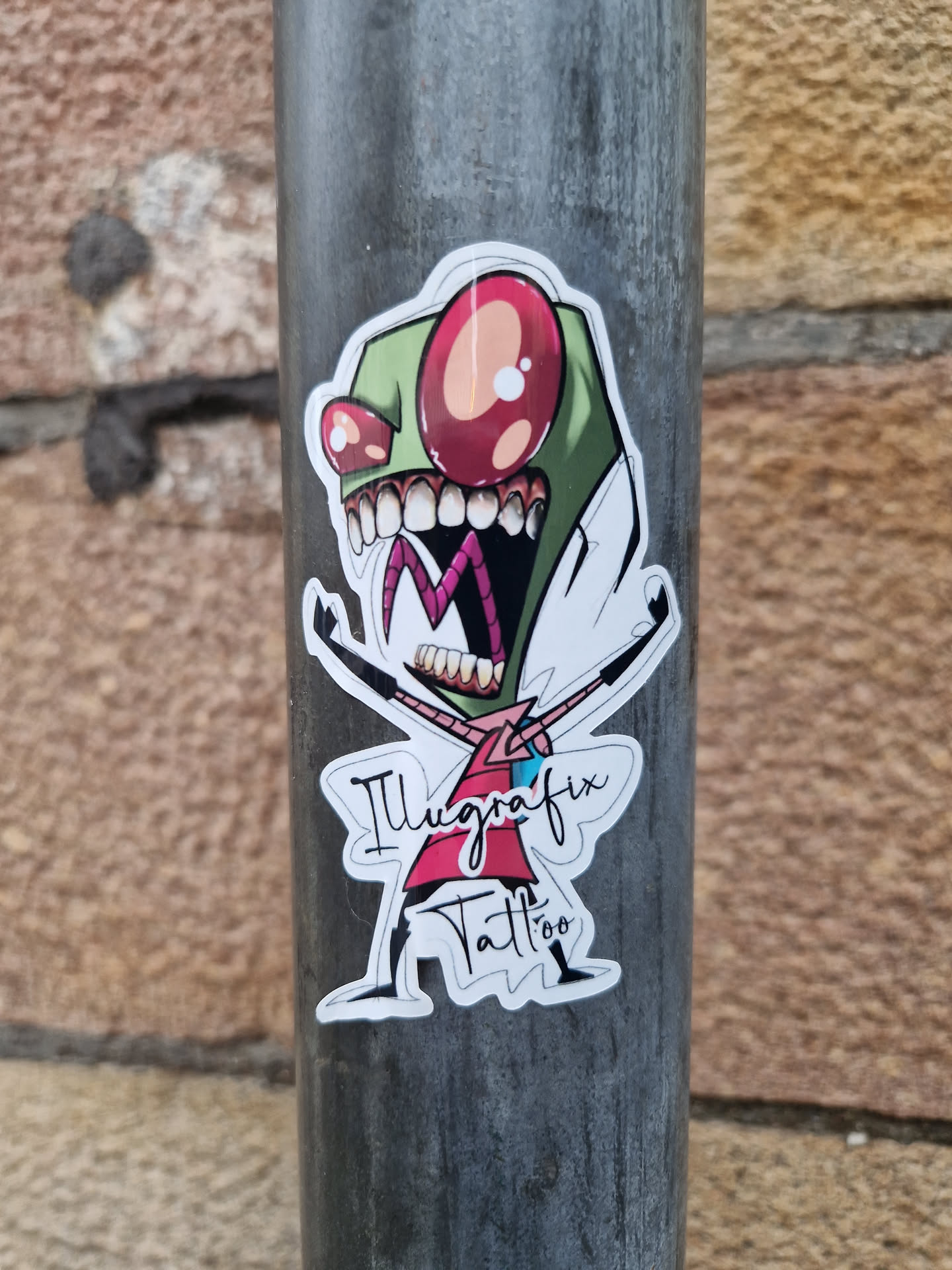 A sticker featuring Invader Zim in a vibrant, stylized design. The artwork is by Illgrafix Tattoo. The character is depicted with his signature wide eyes and menacing grin, arms outstretched in a playful yet slightly menacing pose.  The sticker is adhered to a metal pole.