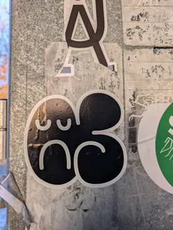 This sticker features a simple black design shaped like a deformed flower or blob with a cute, slightly sad face.  The style is minimalist and cartoonish.