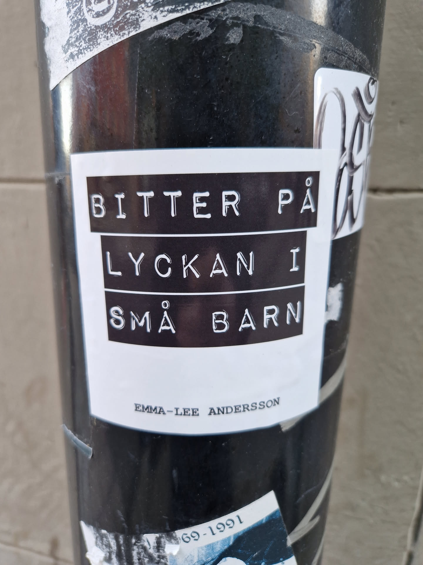 A minimalist sticker with the text "Bitter på lyckan i små barn" in a typewriter-style font, followed by the artist's name, Emma-Lee Andersson. The sticker has a black and white color scheme, and its design is simple yet striking.