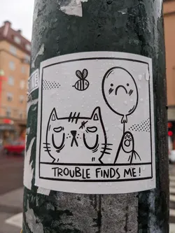 A black and white sticker featuring a cartoon cat, a sad balloon, and a bee. The text 'Trouble finds me!' is written at the bottom. The sticker is wet from the rain.
