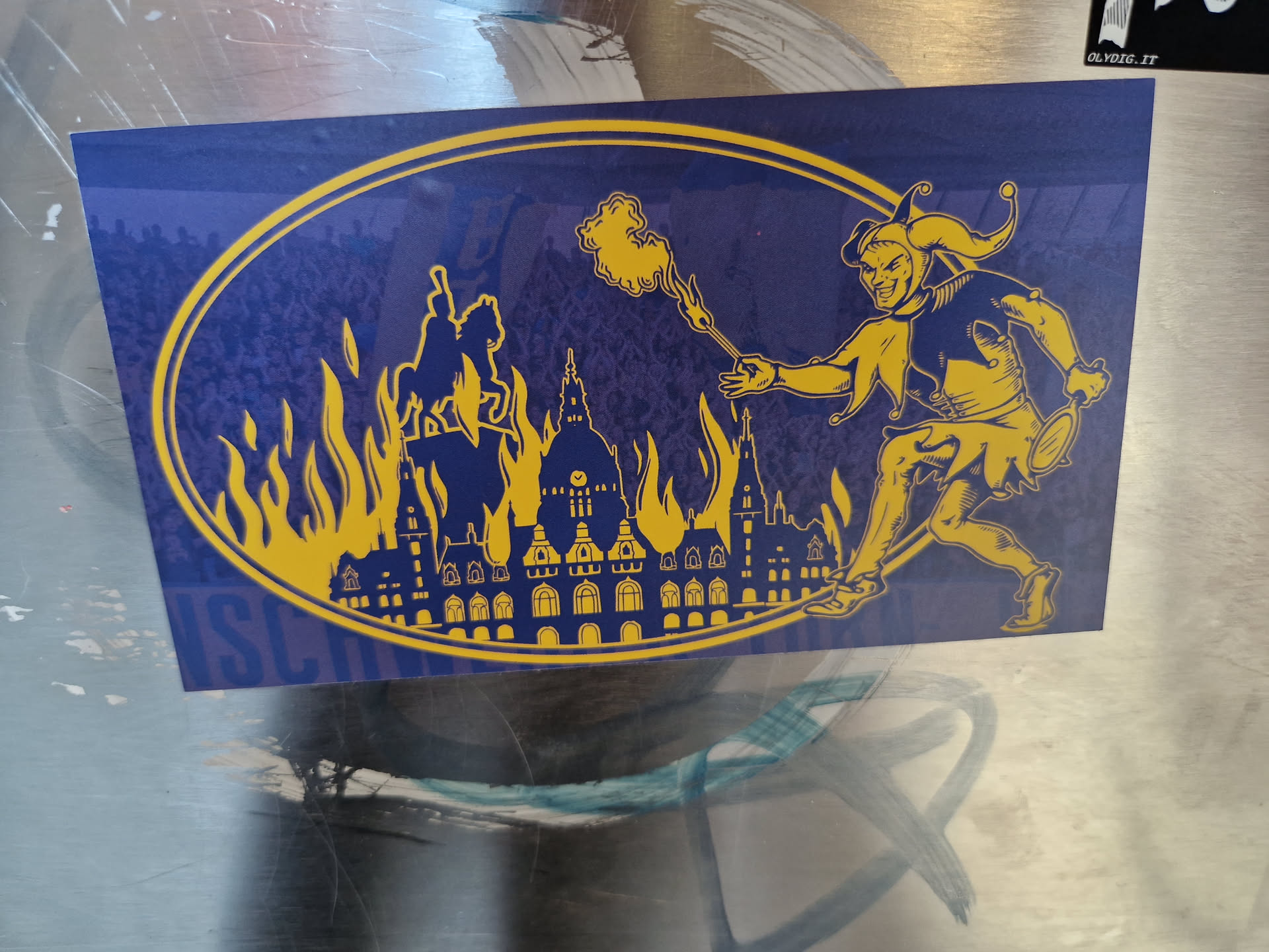 Oval sticker featuring a dark blue background with a yellow jester figure setting fire to a city in silhouette. The jester is depicted in a dynamic pose, wielding a flaming torch. The burning city appears stylized and detailed, with several buildings shown engulfed in flames. The overall style is bold and graphic, reminiscent of historical prints or illustrations.