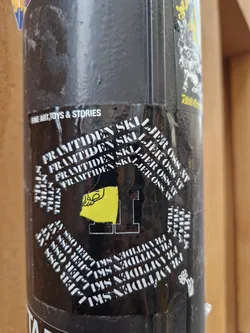 A black and yellow sticker with the text "FRAMTIDEN SKI LJER OSS AT" repeated multiple times in a circular pattern.  There is also a yellow graphic which seems to be a stylized flag or emblem. The text "FINE ART, TOYS & STORIES" is visible on another sticker above it.