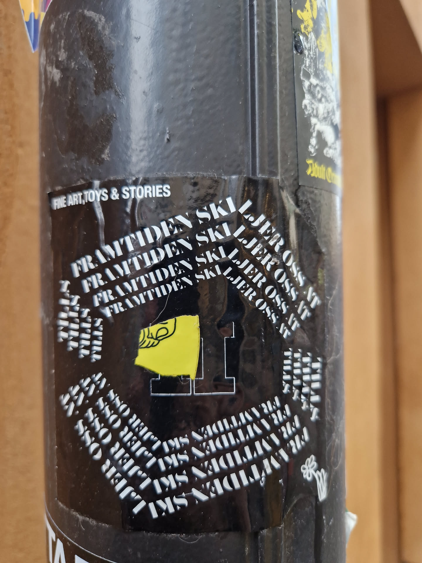 A black and yellow sticker with the text "FRAMTIDEN SKI LJER OSS AT" repeated multiple times in a circular pattern.  There is also a yellow graphic which seems to be a stylized flag or emblem. The text "FINE ART, TOYS & STORIES" is visible on another sticker above it.