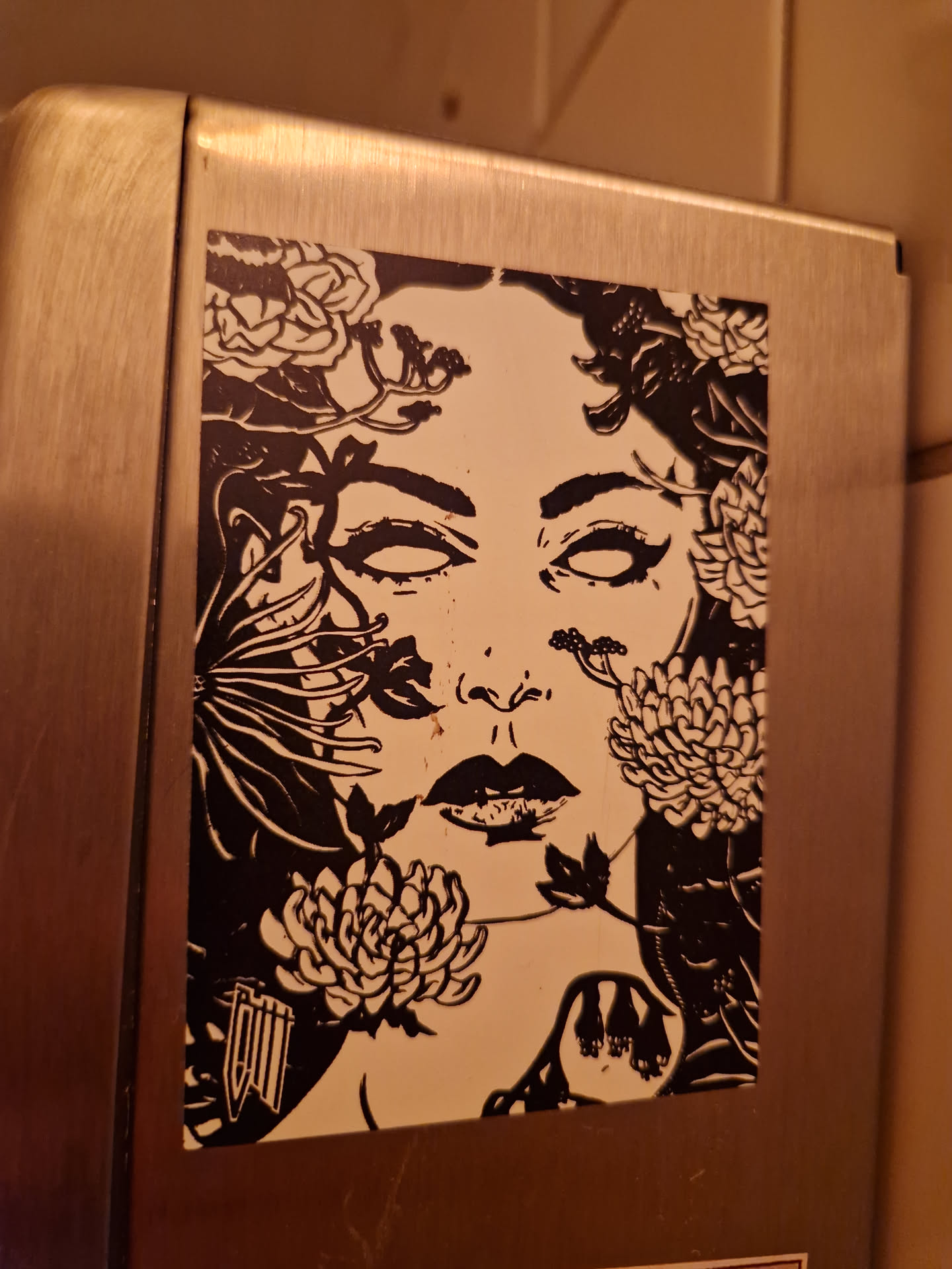 A sticker featuring a black and white illustration of a woman's face surrounded by flowers and plants. The style is reminiscent of botanical illustrations with dark outlines and detailed shading. The woman's expression is serene, and the flowers add a delicate yet dramatic touch to the piece. It's a visually striking image that would make a great addition to any collection.
