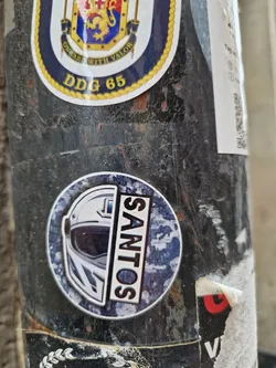 Round sticker featuring the word \"SANTOS\" in bold white letters on a dark blue background with a camouflage pattern.  Half of a white motorcycle helmet graphic is visible on the left side of the sticker.