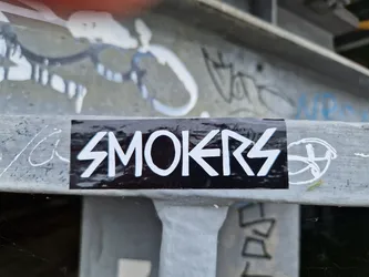 A black and white sticker with the word SMOKERS in a stylized font. The sticker is affixed to a metal surface with graffiti in the background.