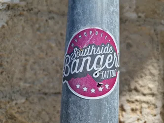 A worn pink and grey round sticker with the text 'Southside Bangers Tattoo' in a stylized font.  The sticker is affixed to a metallic pole.  There are five stars at the bottom of the sticker design. The top of the sticker has 'Monopoli' in a smaller font.