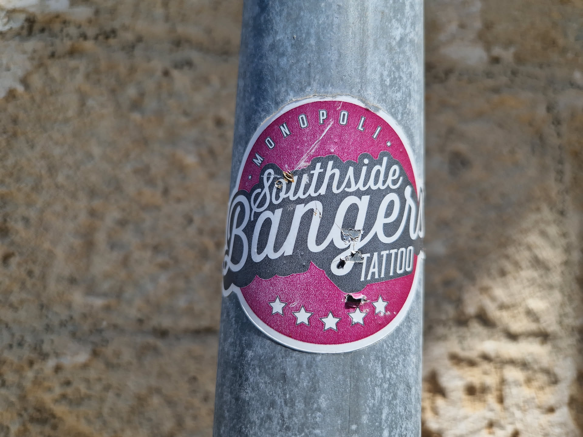 A worn pink and grey round sticker with the text 'Southside Bangers Tattoo' in a stylized font.  The sticker is affixed to a metallic pole.  There are five stars at the bottom of the sticker design. The top of the sticker has 'Monopoli' in a smaller font.