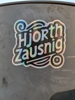 The sticker features a holographic effect, giving it a shimmering appearance. The text "Hjorth Zausnig" is prominently displayed in a stylized font. Below the text is a graphic of a stylized leaf design with a circle at the end, adding a whimsical touch.