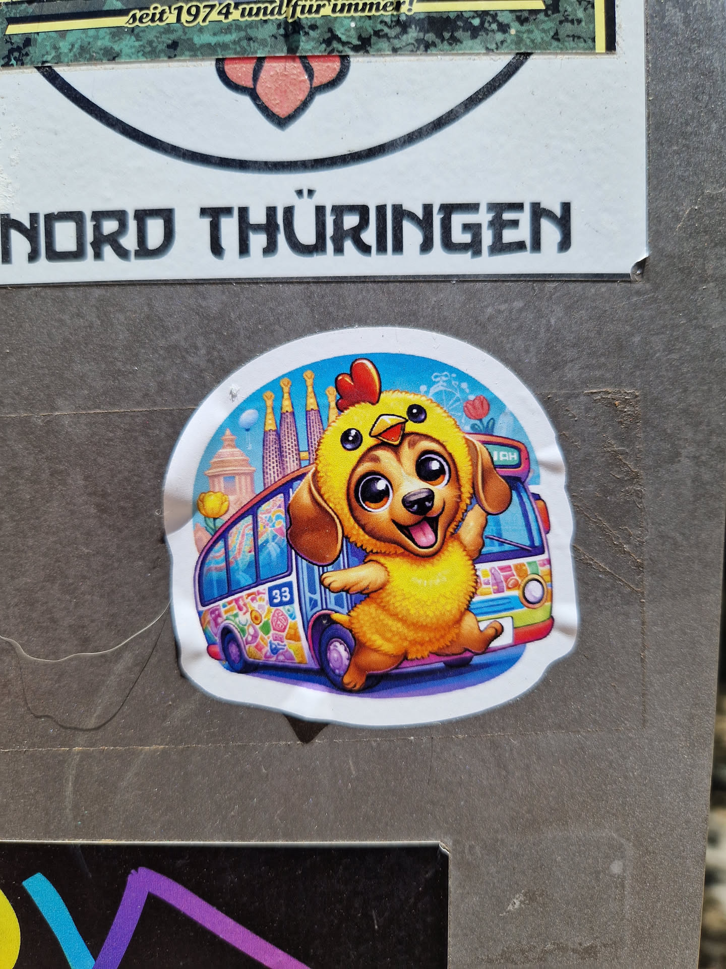 A sticker featuring an illustration of a dog wearing a chicken costume, standing on a colorful psychedelic bus.  The background includes what looks like a stylized cityscape or landmark.