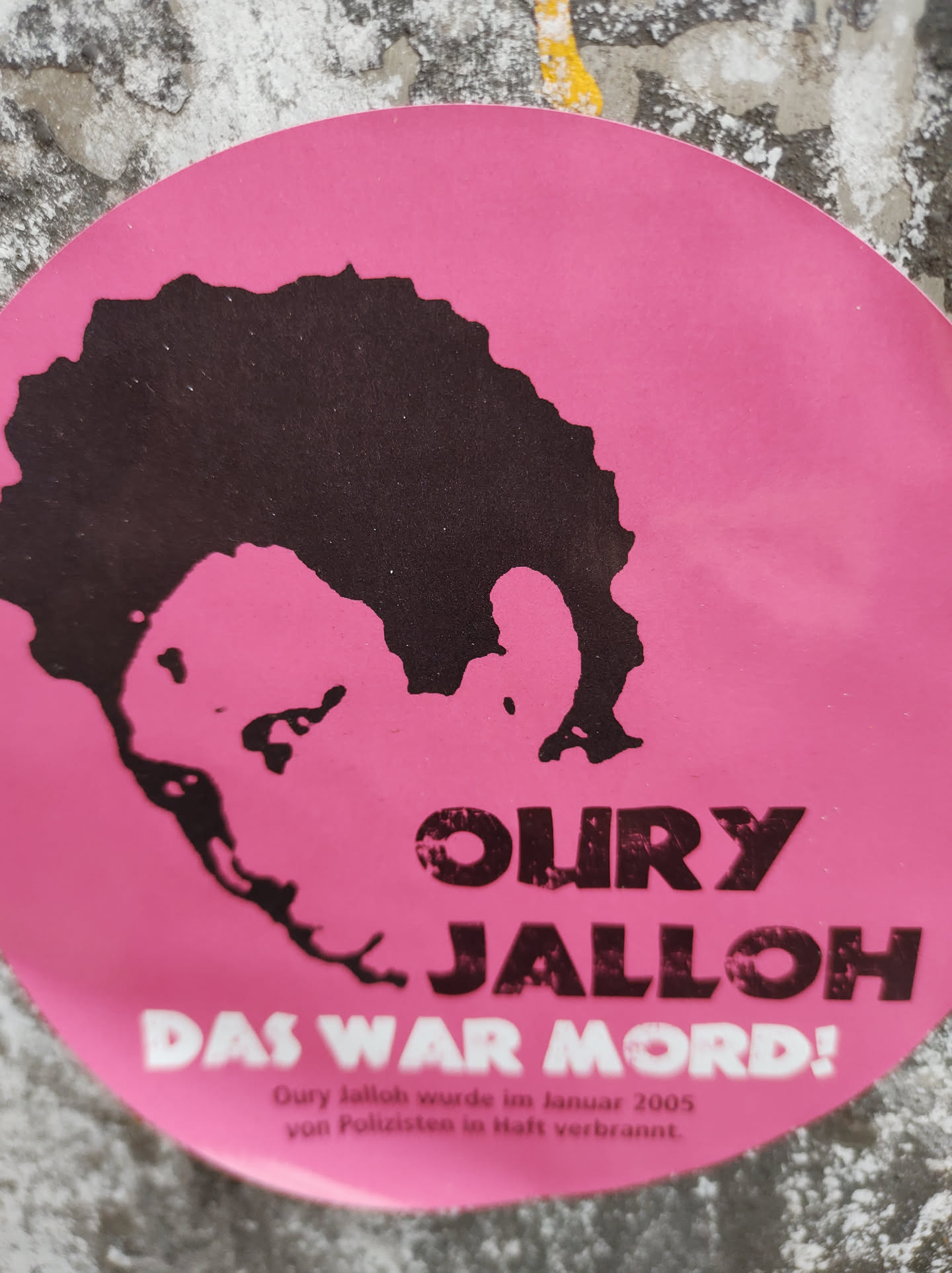 Pink round sticker with a black silhouette of a person's head and shoulders.  Text on the sticker reads "OURY JALLOH" in large letters and "DAS WAR MORD!" in smaller letters. Below, smaller text states that Oury Jalloh was burned to death by police in January 2005. The design appears to be a memorial or protest graphic. 