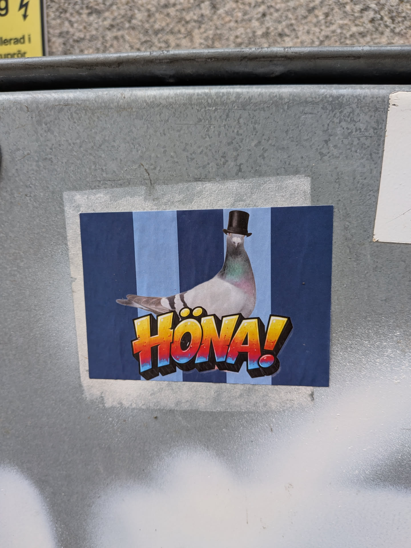 A rectangular sticker depicting a realistic pigeon wearing a small black top hat. The background consists of vertical stripes in alternating shades of blue. In the foreground, the Swedish word 'HÖNA!' is written in a bold, 3D graffiti-style font with a yellow, orange, and red gradient, outlined in black and blue.