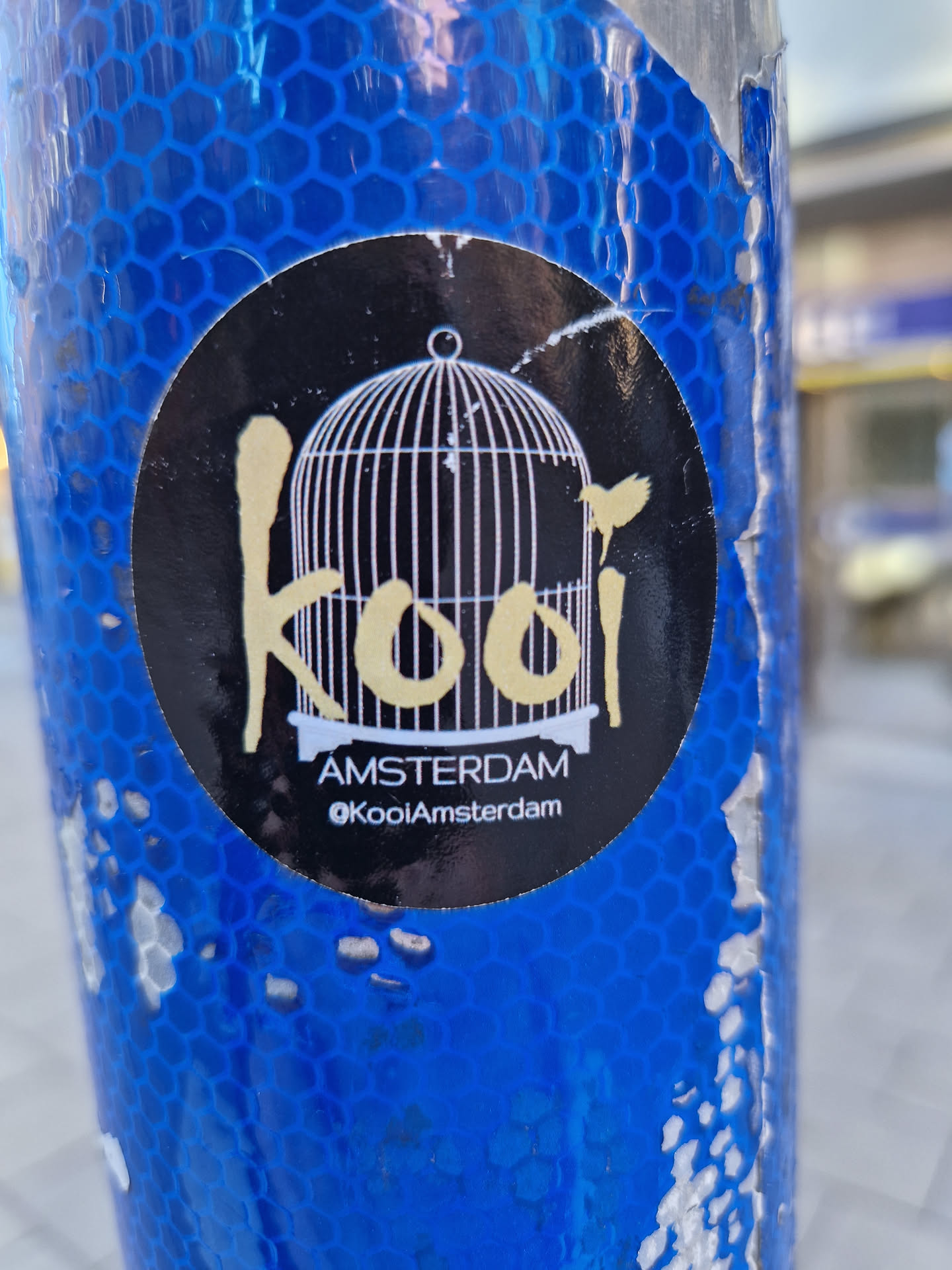 A round black sticker with a stylized text "Kooi" (cage in Dutch) in a yellowish color,  featuring a birdcage graphic and a small bird. Below the text, it says "AMSTERDAM" and "@KooiAmsterdam". The sticker appears to be placed on a textured, blue cylindrical object.