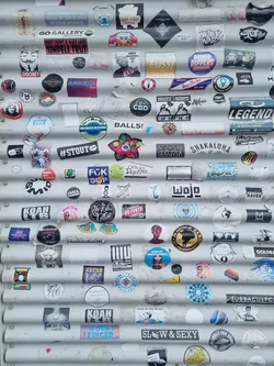 A corrugated metal surface is covered in a variety of stickers, creating a vibrant and layered composition.  Many stickers feature text, logos and abstract designs. The stickers show diverse themes and styles, reflecting a variety of interests and aesthetics.