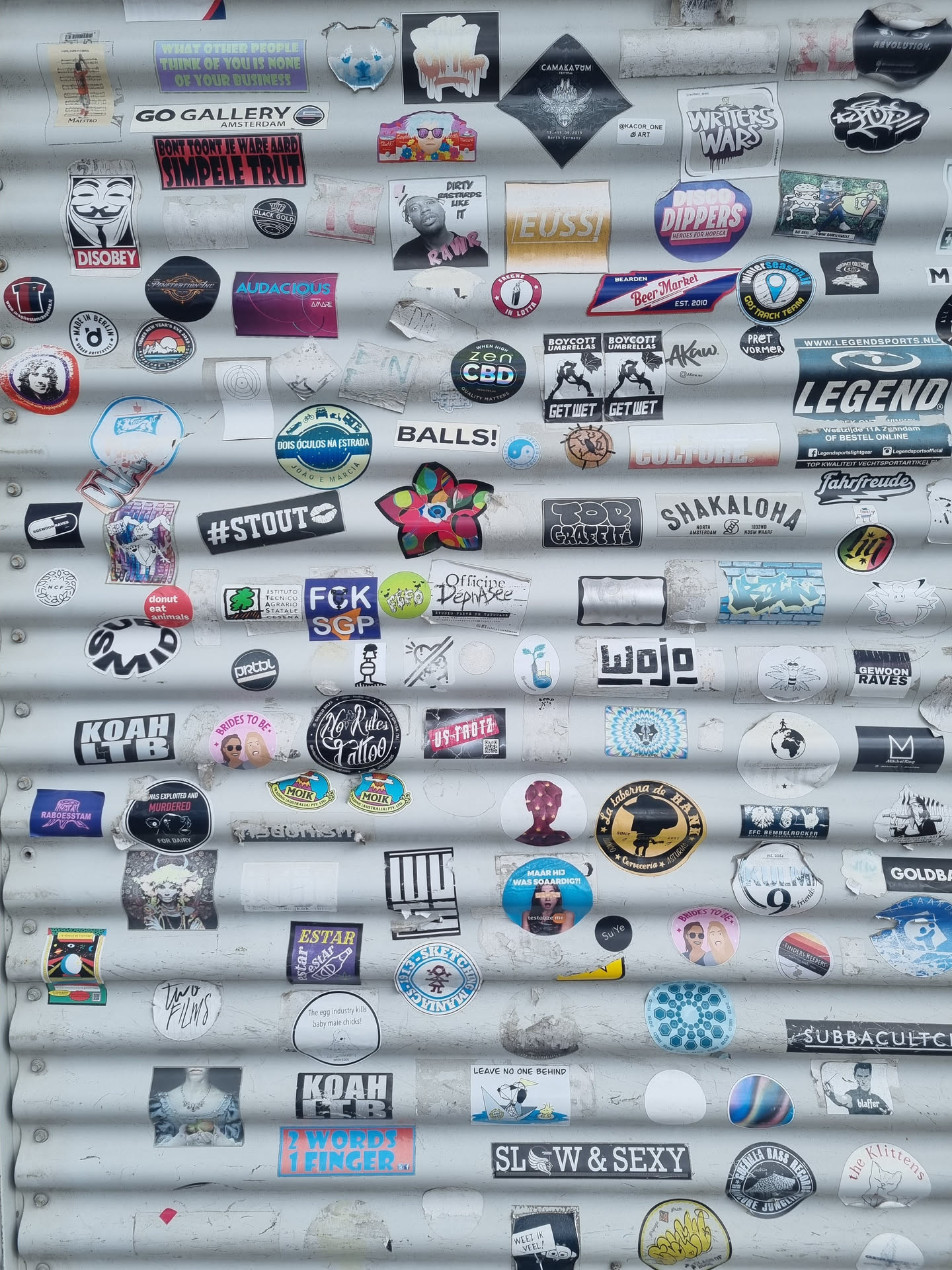 A corrugated metal surface is covered in a variety of stickers, creating a vibrant and layered composition.  Many stickers feature text, logos and abstract designs. The stickers show diverse themes and styles, reflecting a variety of interests and aesthetics.
