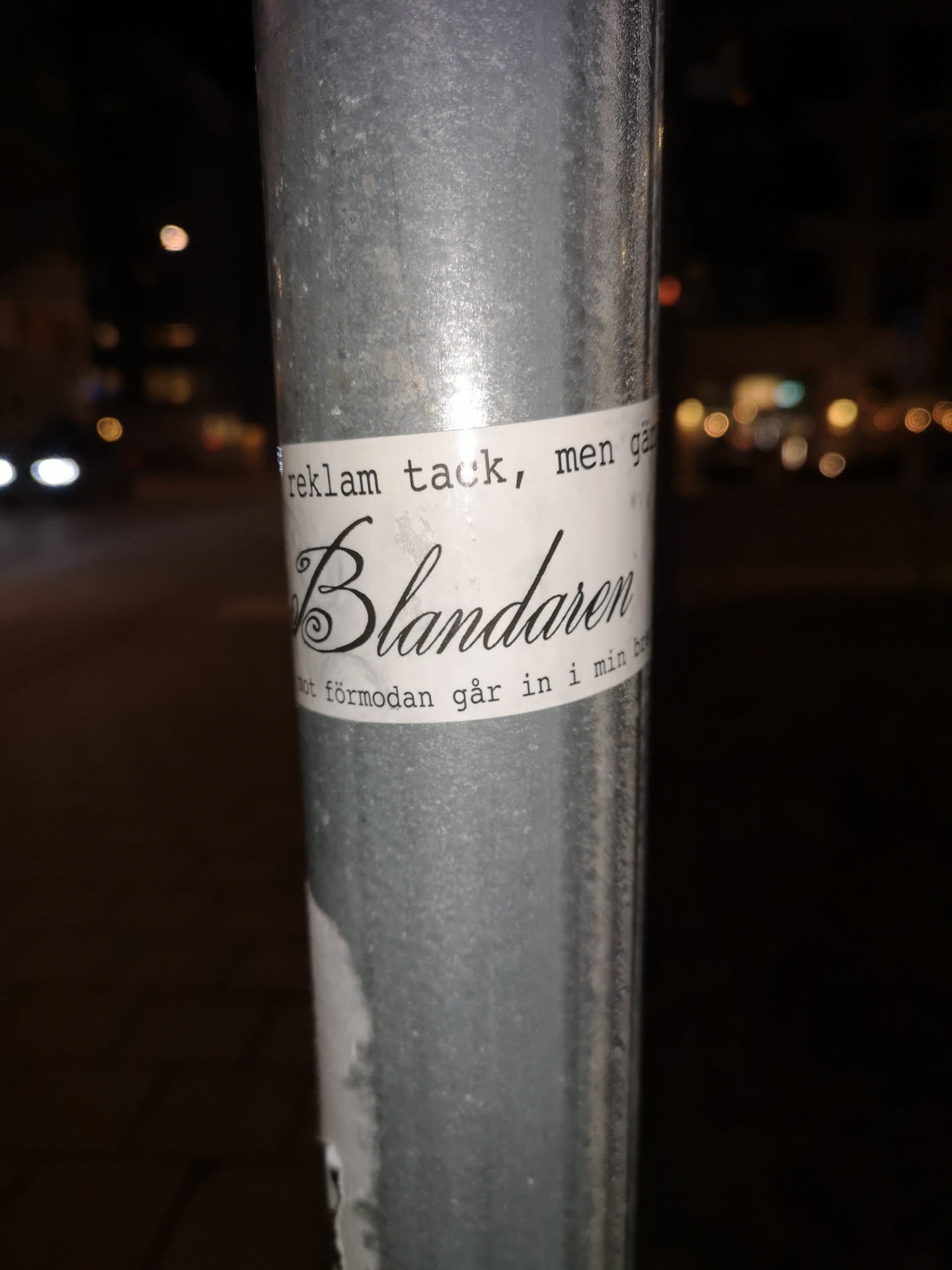 A white rectangular sticker is affixed to a textured, galvanized metal pole at night. The sticker features black text arranged in three lines. The top line, 'reklam tack, men gå', and the bottom line, 'mot förmodan går in i min bo', are in a simple, serif-like font. The central and most prominent word, 'Blandaren', is rendered in an elaborate, elegant calligraphic script with a large, decorative initial 'B'. The sticker shows minor wear, with a slight curl at its bottom edge. The background is blurred, showing distant streetlights and vehicle lights.