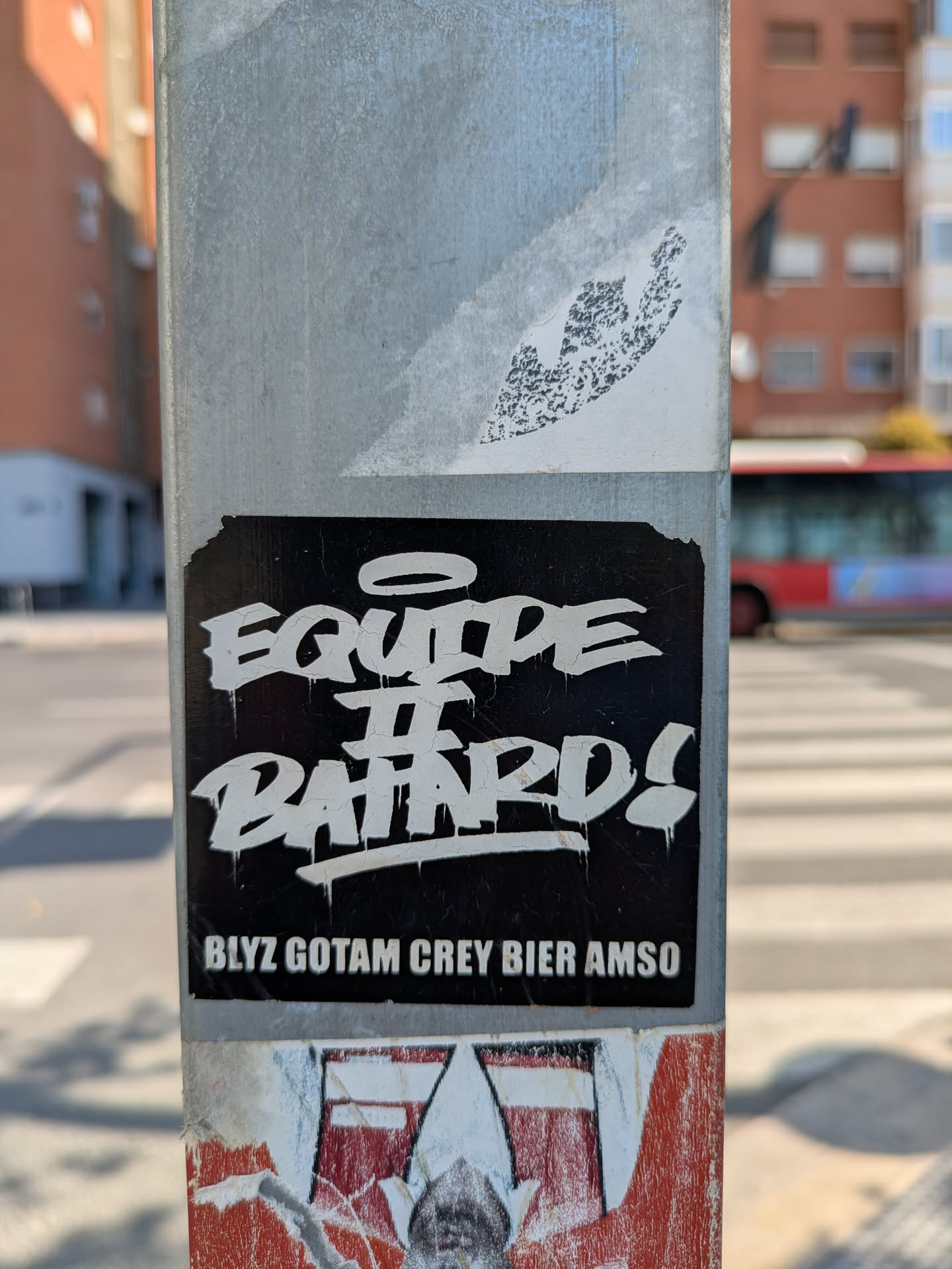 A black and white sticker on a metal pole reads "Equipe II Batards!". Below that, the text "BLY2 GOTAM CREY BIER AMSO" is displayed. The text is white on a black background. The sticker appears somewhat aged.