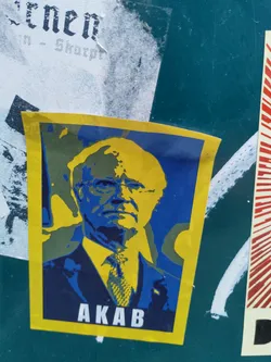 A yellow and blue sticker with a stylized portrait of a man, possibly a political figure. The text 'AKAB' is visible at the bottom.  The style is reminiscent of protest art.