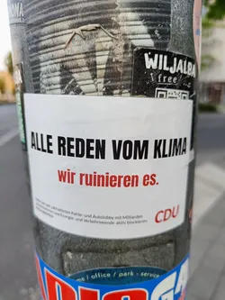 A sticker on a pole with the text "Alle reden vom Klima, wir ruinieren es." (Everyone talks about the climate, we are ruining it.)  The sticker also contains the CDU logo. 
