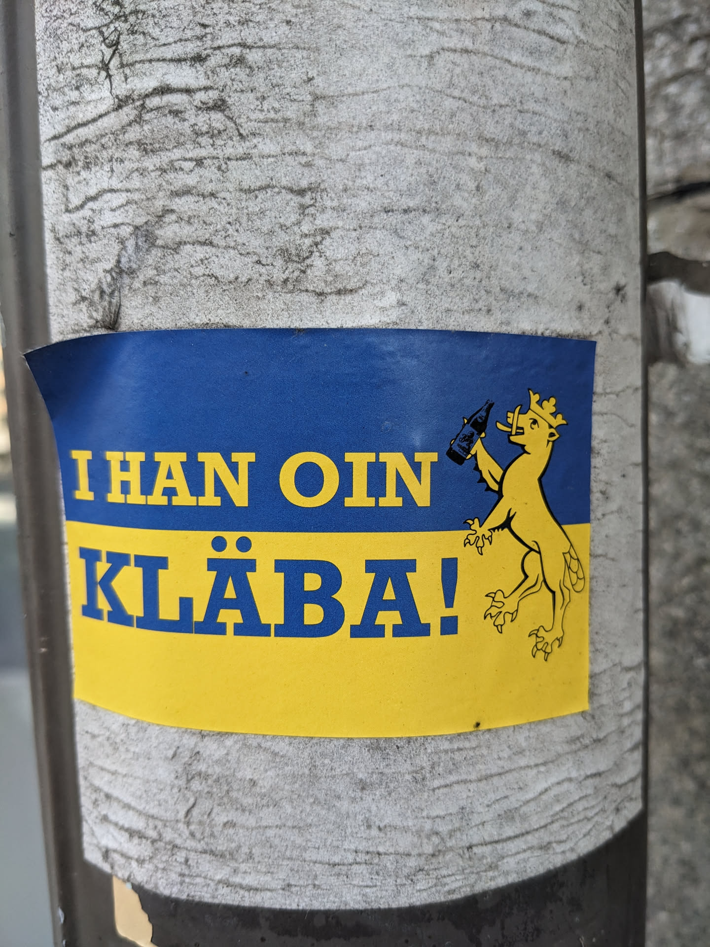 A yellow and blue sticker featuring a crowned lion drinking from a bottle and the text "I han oin kläba!" in blue lettering. The phrase is a playful take on the Swedish language.