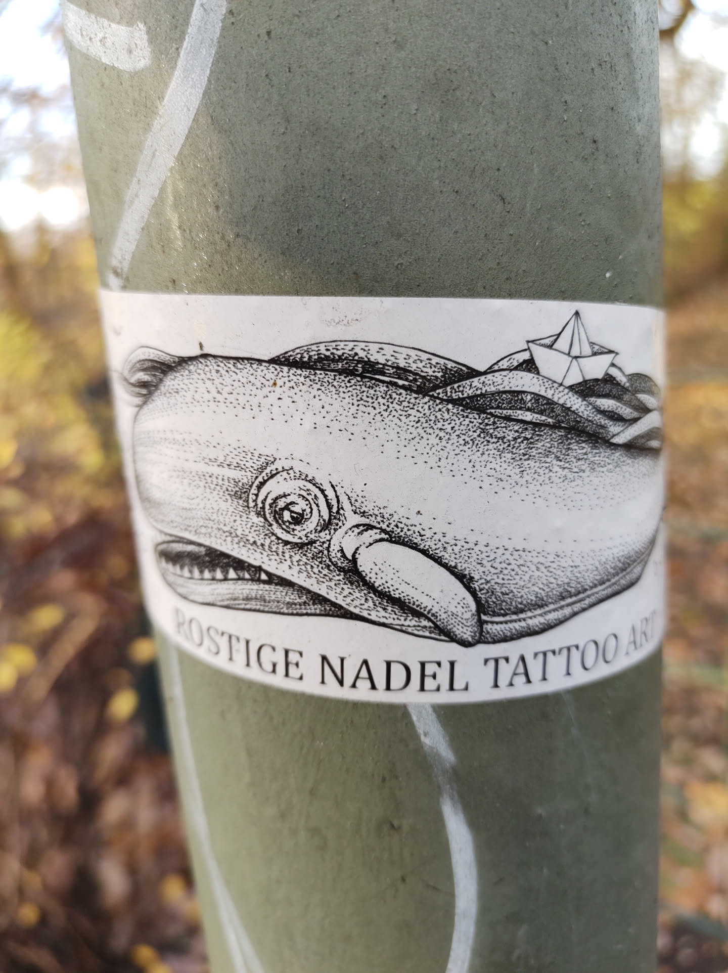 Black and white illustration of a whale with a small paper boat on wavy water. The sticker has the text "Rostgie Nadel Tattoo Art" at the bottom.
