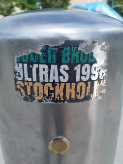 A worn sticker on a metal pole. The sticker features the text 'Ultras 1999 Stockholm' in a distressed style with some parts of the text missing or damaged. The colors are mainly black, white, teal, and gold.