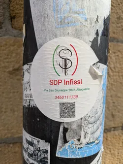 A round SDP Infissi sticker is pasted on a pole. The logo features a stylized key encircled by green and red lines, reminiscent of the Italian flag. Below the logo is the company name "SDP Infissi," followed by their address "Via San Giuseppe 20/2, Altopascio" and a phone number, "3460111739". A QR code is located at the bottom.