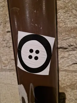 A simple, black and white sticker depicting a button with four holes. It's affixed to a brown cylindrical pole against a stone wall.  The design is clean and modern.