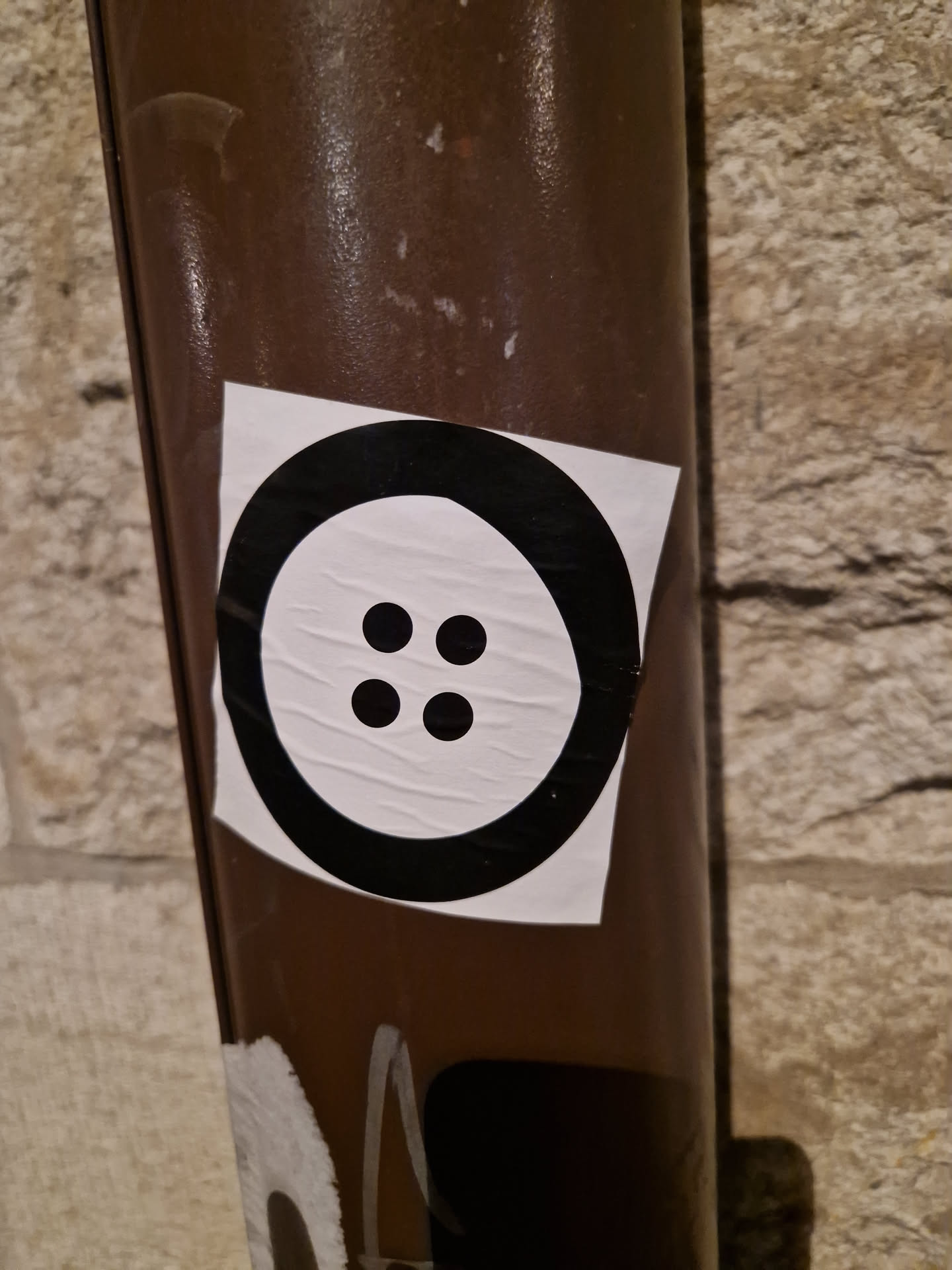 A simple, black and white sticker depicting a button with four holes. It's affixed to a brown cylindrical pole against a stone wall.  The design is clean and modern.