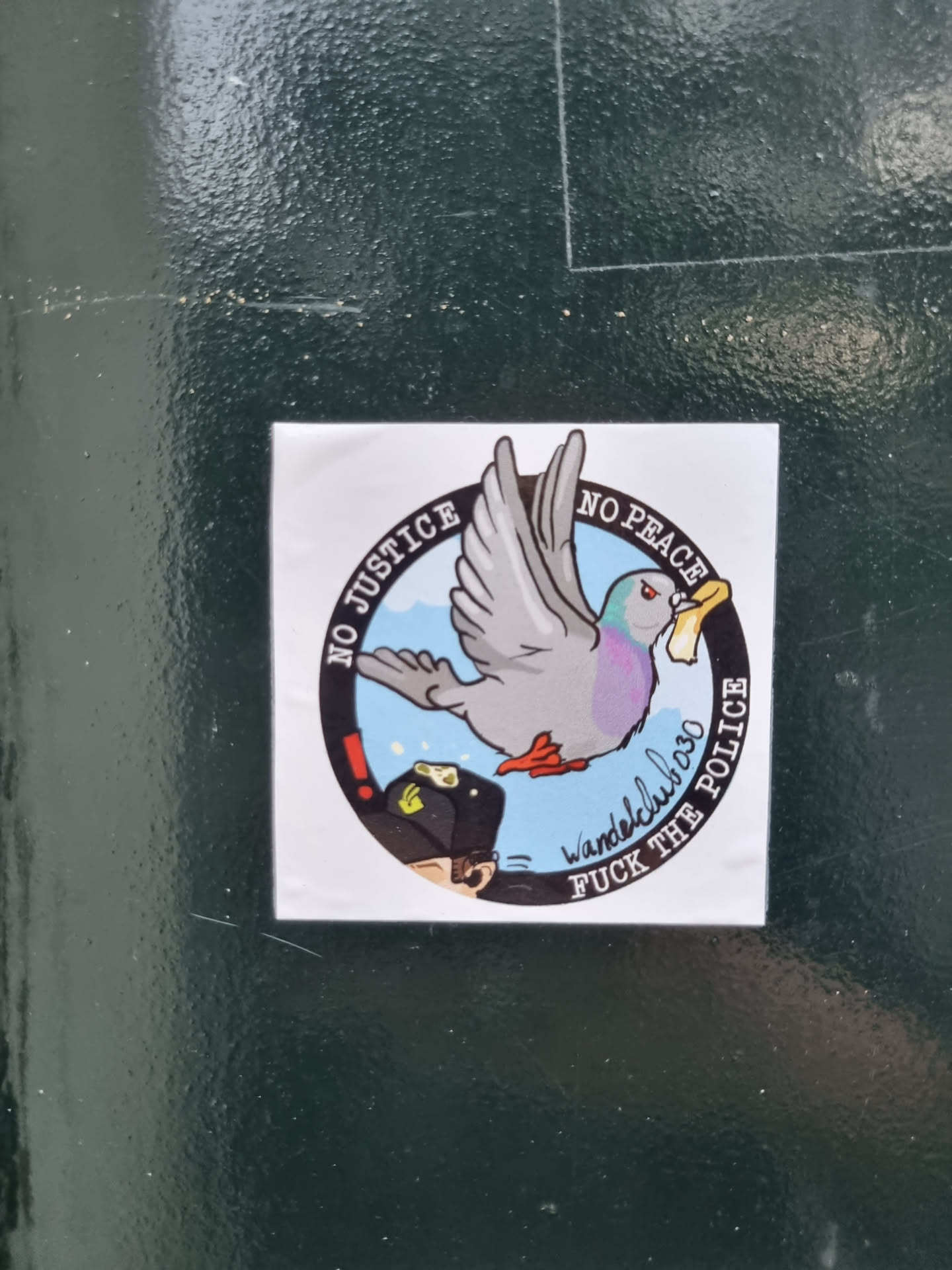 A sticker with an illustration of a pigeon carrying a piece of paper in its beak. The sticker has a circular design with the text "No Justice No Peace" written on the upper part of the circle and "Fuck The Police" on the lower part.  In the center is a cartoon pigeon flying.  The artist's signature, "wandelcluboso", is also included on the sticker.