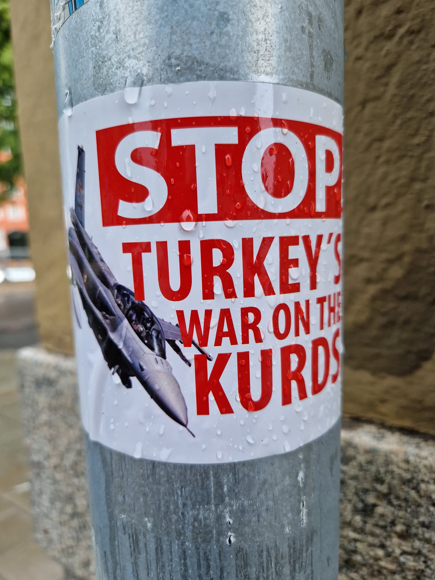 A sticker depicting a fighter jet and the text "STOP TURKEY'S WAR ON THE KURDS" in red and white. The sticker is affixed to a metal pole outdoors and shows water droplets.