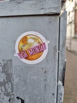 Round sticker with a golden globe and the text Hash Montana written in a purple neon-like style. The globe is stylized and has continents outlined in darker gold.